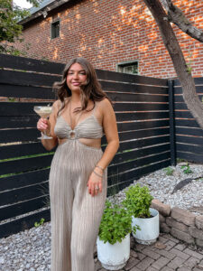 Tristen in a gold jumpsuit standing on her brick patio holding a lemon drop martini