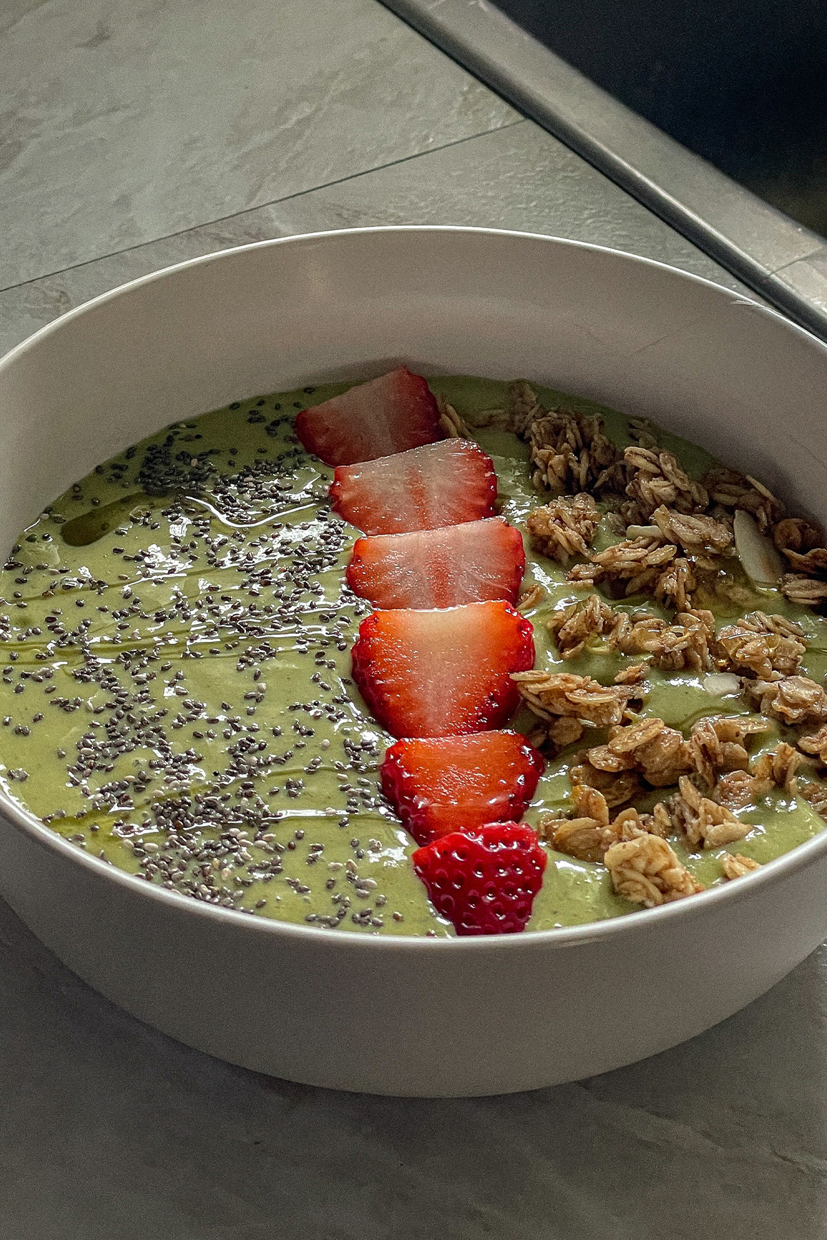 Tropical green smoothie bowl topped with granola, sliced strawberries, chia seeds and honey