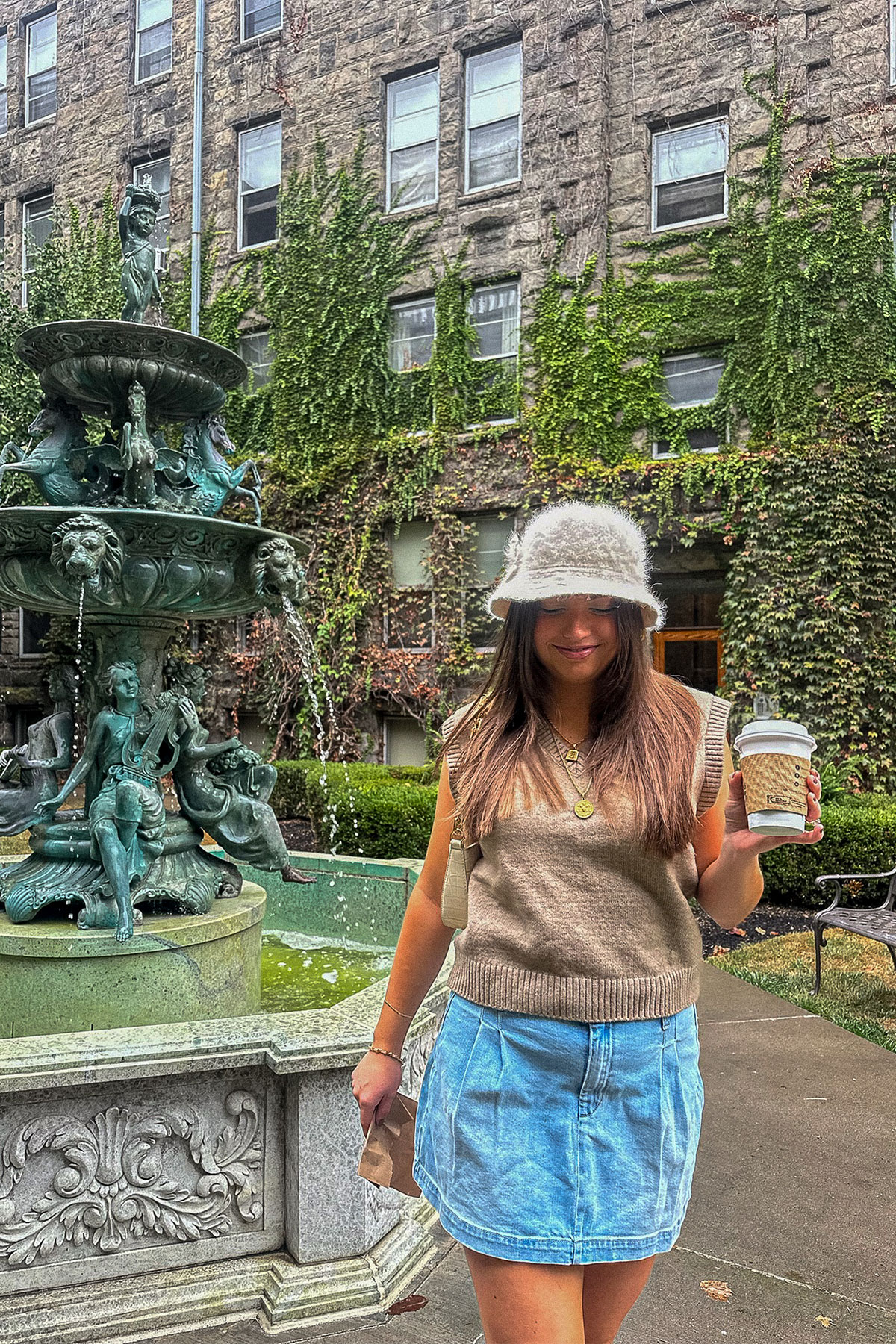 Tristen wearing a pleated denim skirt, beige sweater vest and white furry bucket hat in a vine-covered courtyard