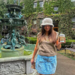 Tristen wearing a pleated denim skirt, beige sweater vest and white furry bucket hat in a vine-covered courtyard