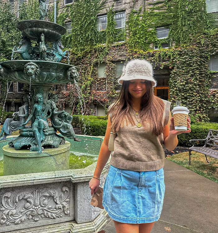 Tristen wearing a pleated denim skirt, beige sweater vest and white furry bucket hat in a vine-covered courtyard